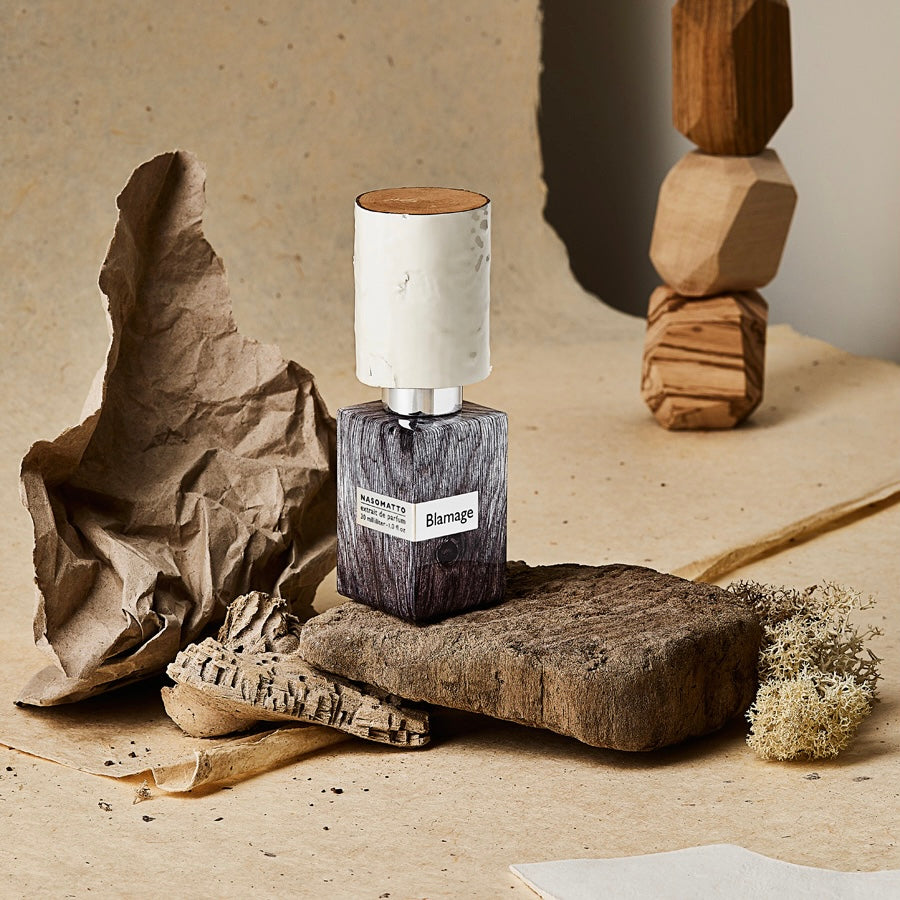 Perfume bottle labeled 'Blamage' on a textured surface with natural elements.